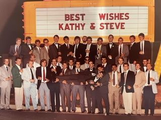 A large group in jackets and ties under a marquee reading Best Wishes Kathy & Steve