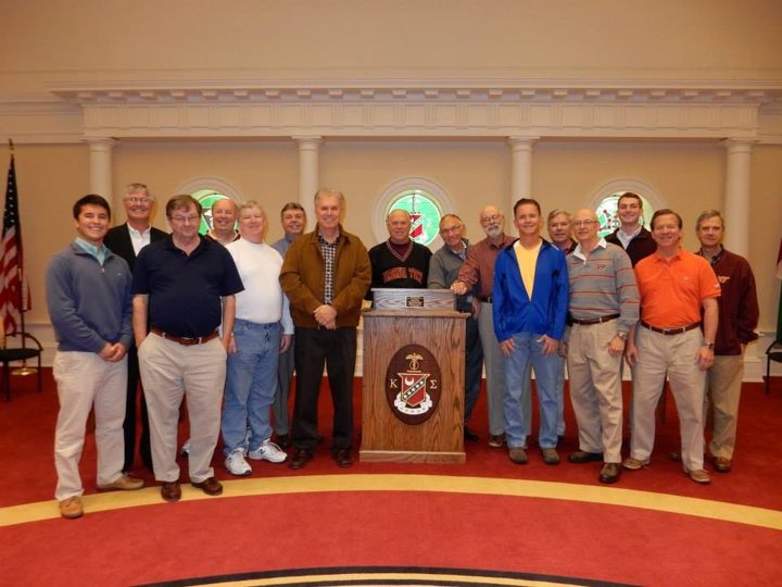 A large group of brothers standing around a lectern with the Kappa Sigma crest in a domed hall